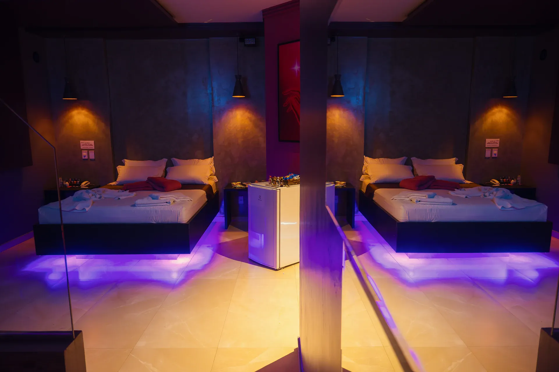 Suíte Hidro Plus com cama refletida no espelho e iluminação violeta e âmbar
