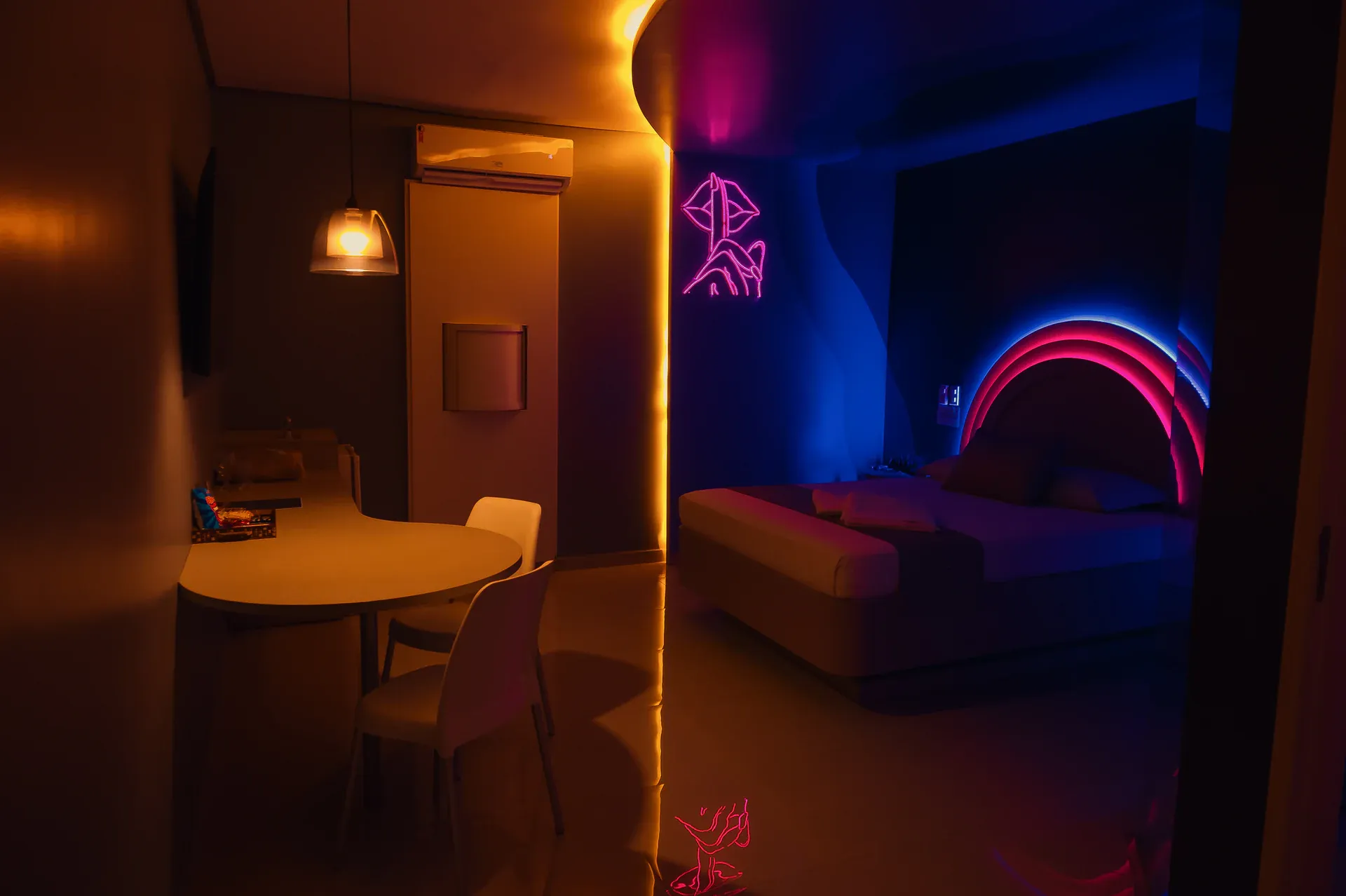 Suíte Luxo com cama em neon azul e rosa e mesa sob luz âmbar