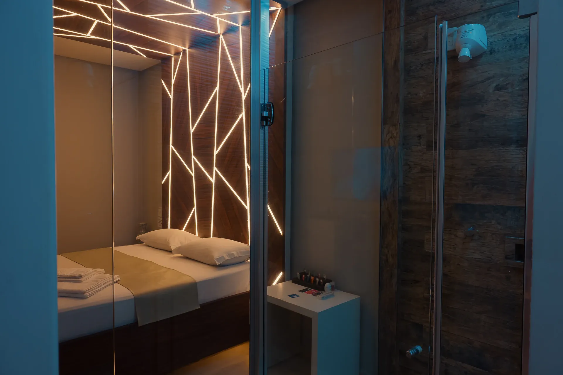 Cama com painel geométrico iluminado vista através do box de vidro