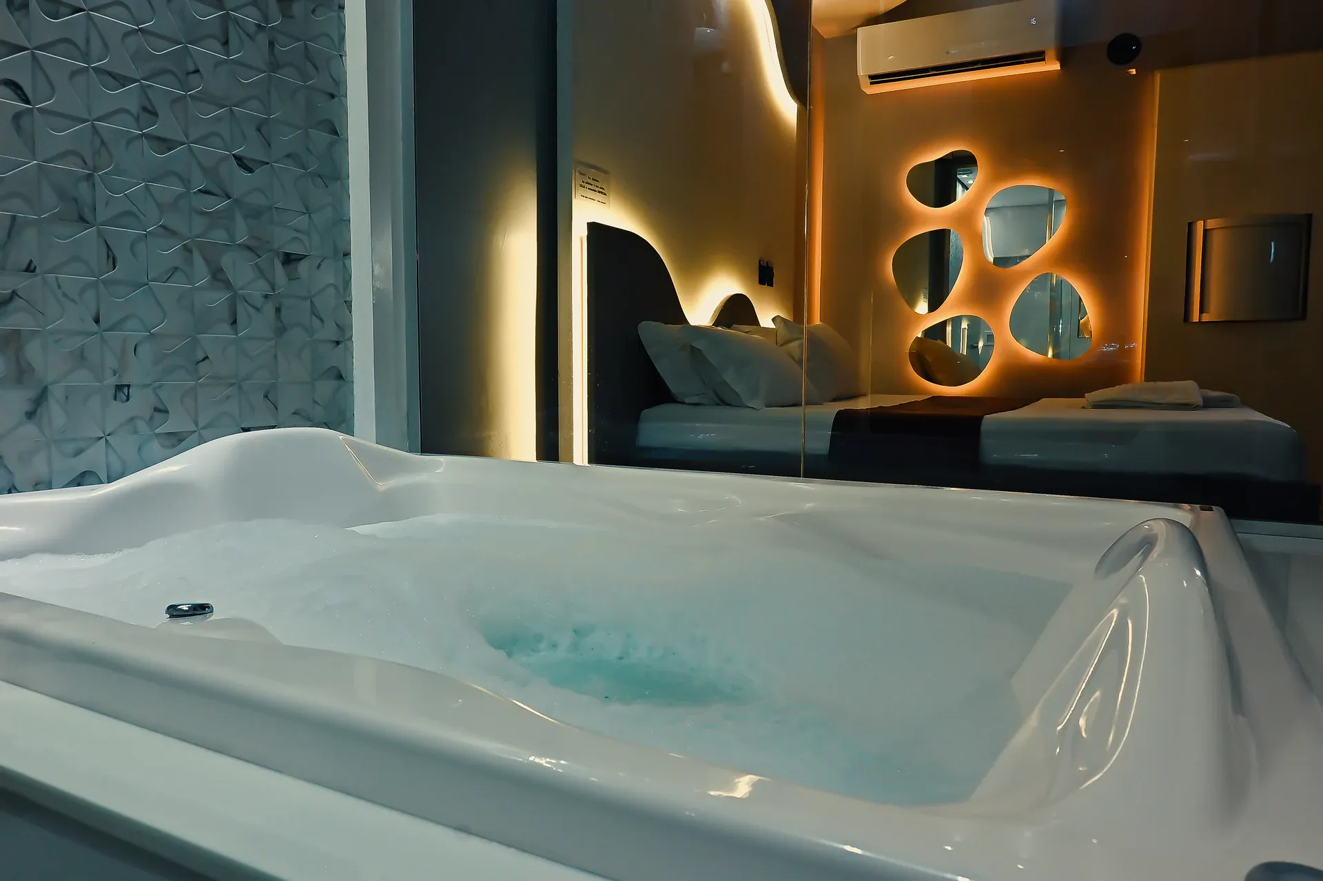 Hidromassagem com espuma em primeiro plano e cama iluminada atrás do vidro
