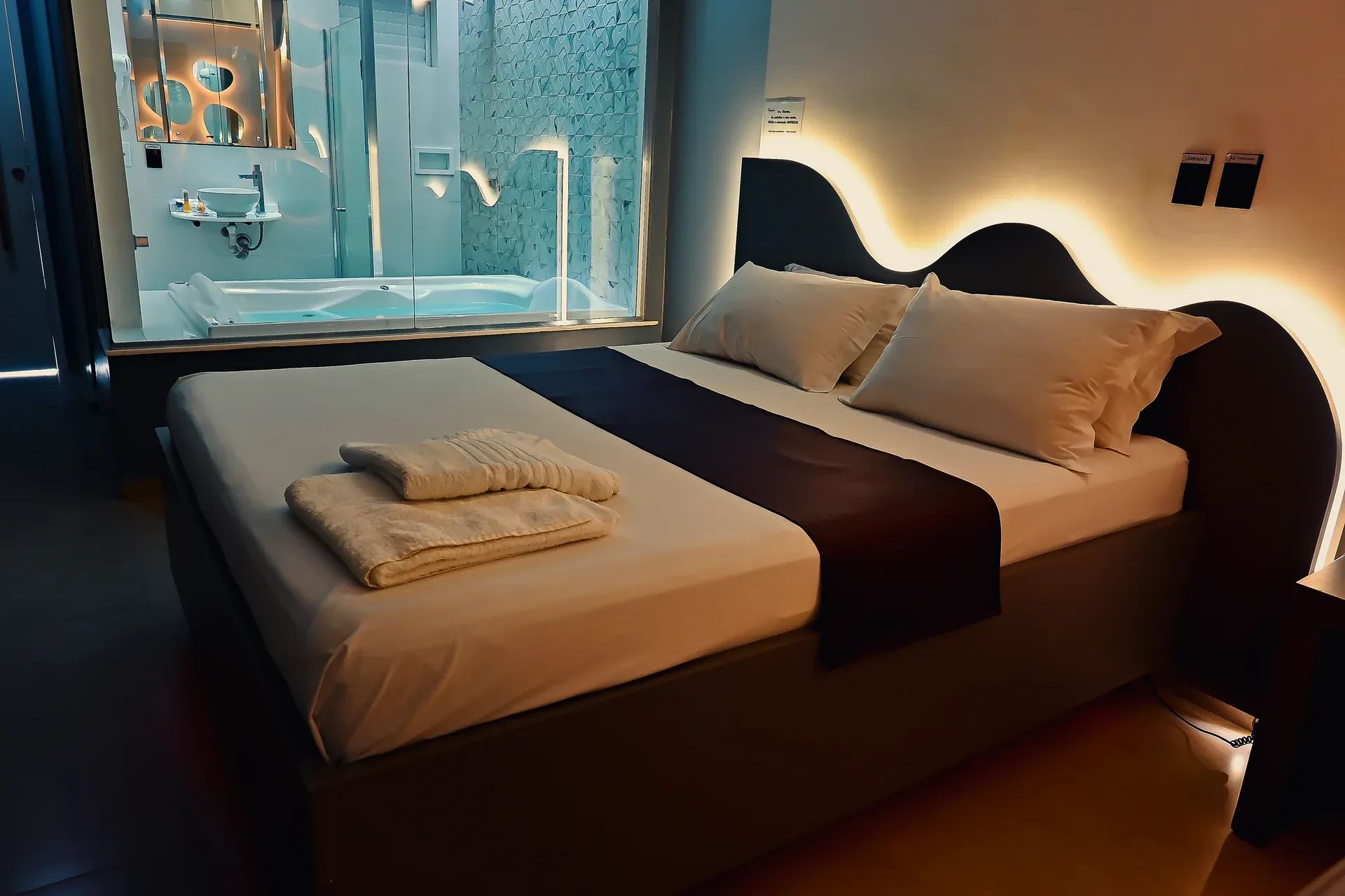 Cama com cabeceira ondulada iluminada e hidromassagem visível atrás do vidro
